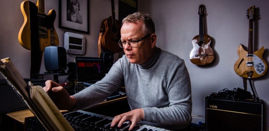 A Caucasian male wearing glasses and a grey jumper plays piano in a room with guitars hanging on the wall.